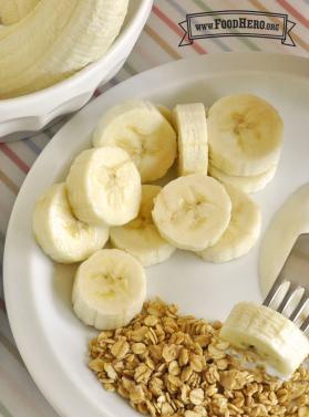 A fork is used to dip chunks of banana into yogurt and then into granola for a simple snack.