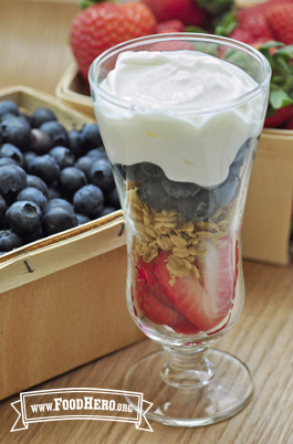 A sundae glass filled with layers of sliced strawberries, granola, blueberries and creamy yogurt.