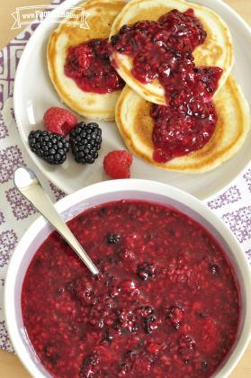 A serving bowl of Any Berry Sauce is shown with a plate of 3 pancakes that have sauce spread on top and a few fresh berries on the side.