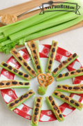 Celery sticks spread with peanut butter and topped with raisins are displayed on a plate in a pinwheel formation. In the middle of the plate is a celery and peanut butter stick with mini pretzels extending out of the peanut butter like the wings of a butterfly.