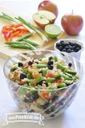 A clear bowl displays a colorful salad of cooked barley, blueberries and crunchy vegetables.