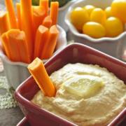 Bowl of hummus with a lemon slice served with carrot sticks, tomatoes, and broccoli.