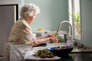 older woman rinsing a carrot and other produce under running water at the kitchen sink