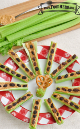 Celery sticks spread with peanut butter and topped with raisins are displayed on a plate in a pinwheel formation. In the middle of the plate is a celery and peanut butter stick with mini pretzels extending out of the peanut butter like the wings of a butterfly.