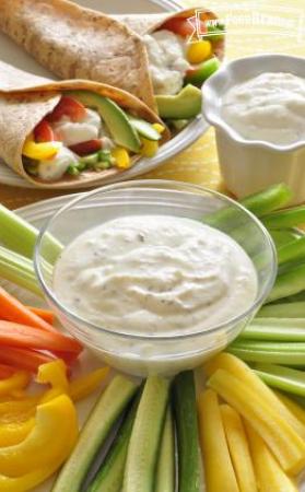 Small bowl of creamy dip with a plate of sliced vegetables and served in a veggie wrap.
