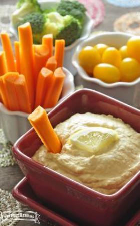 Bowl of hummus with a lemon slice served with carrot sticks, tomatoes, and broccoli.