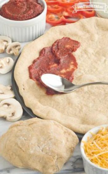 Pizza dough shown with smeared red sauce and pizza toppings.